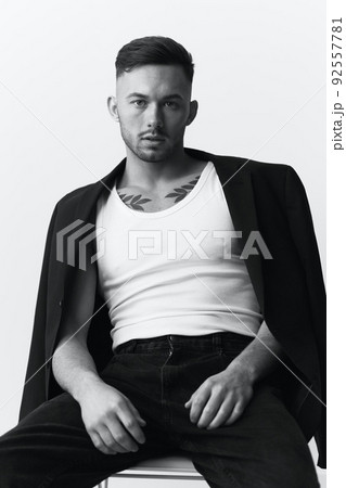 Modelling snapshots. Serious self-confident attractive handsome man in black jacket sitting on chair posing in white studio background. Black and White concept. Copy space 92557781
