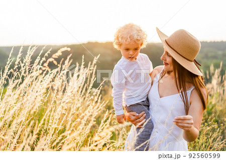 Pretty woman holding her son in her arms while walking in a picturesque field at sunset 92560599