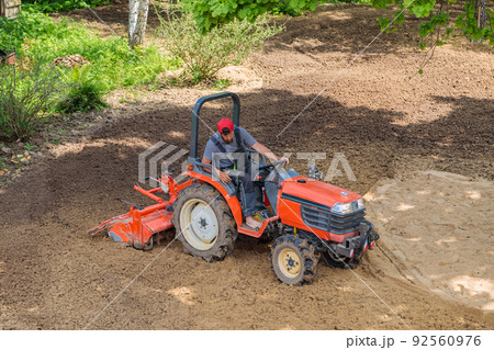 A farmer on a mini tractor loosens the soil for the lawn. Land cultivation, surface leveling A farmer on a mini tractor loosens the soil for the lawn. Land cultivation, surface leveling 92560976