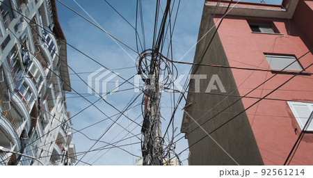 Tangled electricity fiber optic cables in communication systems, against blue sky, residential apartment buildings. Mess in black wires of power supply on electrical pole. Telecommunications problems Tangled electricity fiber optic cables in communication systems, against blue sky, residential apartment buildings. Mess in black wires of power supply on electrical pole. Telecommunications problems 92561214
