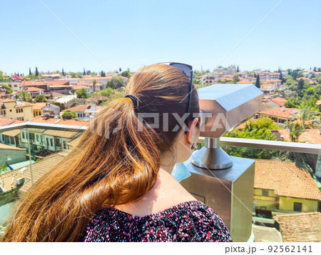binoculars for tourists are installed on the roof. a girl with long, dark hair in a ponytail looks at houses with tiled roofs. woman on a sightseeing tour in a tropical country 92562141