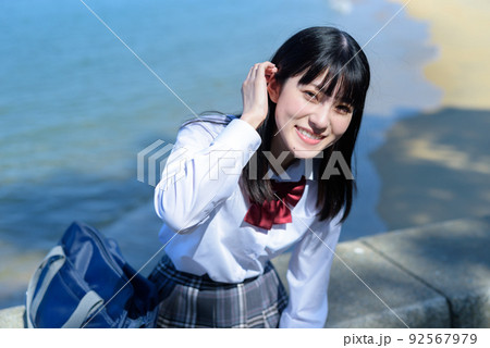 綺麗な海辺の防波堤に座って髪をかき上げながら上目遣いの笑顔でこちらを見る女子高校生　放課後　波打ち際 92567979