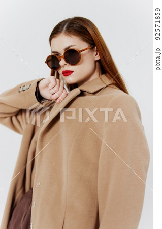 A stylish young woman in glasses and a beige coat poses on a white background in designer clothes at a catalog photo shoot in the studio 92571859