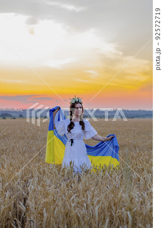 Happy girl in national Ukrainian embroidered shirt with wreath on his head hold large yellow-blue flag against background of spikelets of wheat field. Independence Day. Glory to Ukraine. Patriotism Happy girl in national Ukrainian embroidered shirt with wreath on his head hold large yellow-blue flag against background of spikelets of wheat field. Independence Day. Glory to Ukraine. Patriotism 92572719