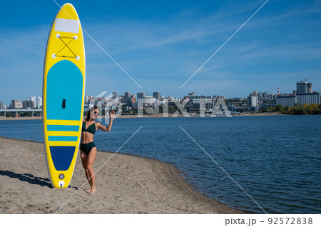Caucasian woman walks along the beach and carries a sup board on the river in the city. Summer sport. 92572838