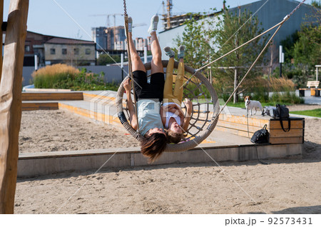 Mom and daughter swing on a round swing. Caucasian woman and little girl have fun on the playground. Mom and daughter swing on a round swing. Caucasian woman and little girl have fun on the playground. 92573431