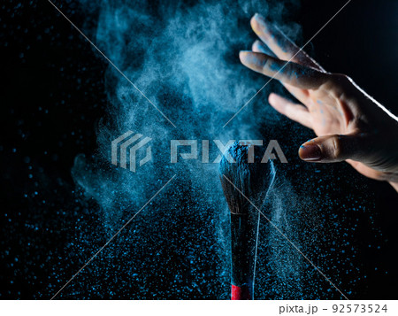 Woman shaking off blue powder from a brush on a black background. Woman shaking off blue powder from a brush on a black background. 92573524