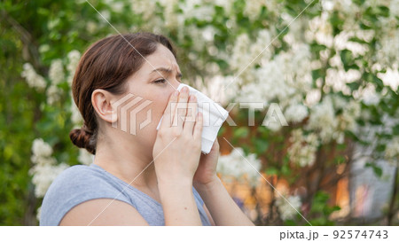 Caucasian woman suffers from allergies and blows her nose into a napkin while walking in the park. Caucasian woman suffers from allergies and blows her nose into a napkin while walking in the park. 92574743