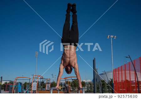Shirtless man doing handstand on parallel bars at sports ground. Shirtless man doing handstand on parallel bars at sports ground. 92575630