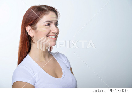 Portrait of a redhead woman with tapes on skin color face for rejuvenation. An alternative way to fight wrinkles. 92575832