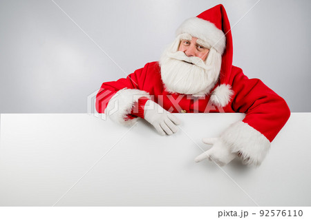 Santa Claus peeks out from behind a white background and points to an empty space with his finger. Merry Christmas.  92576110