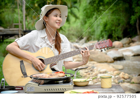 Relaxed and peaceful young Asian female camper traveler singing song and playing guitar 92577505