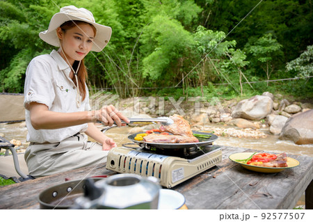 Pretty Asian female traveler preparing her picnic lunch, making a barbecue steak. 92577507