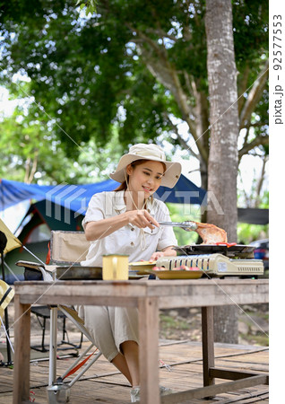Portrait, Asian female camper grill some steaks on a portable outdoor picnic stove at campground. Portrait, Asian female camper grill some steaks on a portable outdoor picnic stove at campground. 92577553