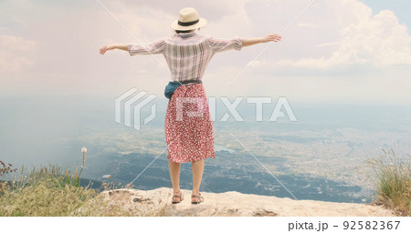 Woman traveler flaps arms like wings on mountain top. Young woman in hat opens arms wide waving like bird's wings over Tira city, Albania. Red dress happy girl blowing wind. Dream, travel concept 92582367