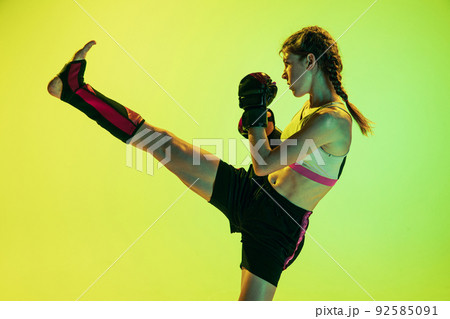 Dynamic portrait of athletic girl, MMA fighter training isolated on gradient yellow-green background in neon. Concept of sport, energy, combat sports 92585091