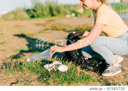 Young female volunteer satisfied with picking up trash, a plastic bottles and coffee cups, clean up beach with a sea. Woman collecting garbage. Environmental ecology pollution concept. Earth Day 92585720