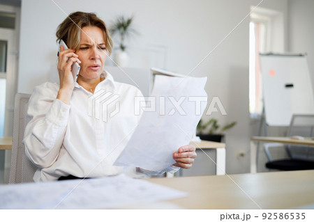 Middle aged business woman with a serious face talking on a smartphone while looking at documents 92586535