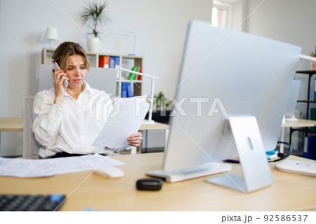 Woman accountant indignantly looks at documents and talks on the smartphone Woman accountant indignantly looks at documents and talks on the smartphone 92586537