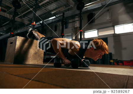 Portrait of young sportive red-headed man doing push up exercises with dumbbells isolated on indoor gym background Portrait of young sportive red-headed man doing push up exercises with dumbbells isolated on indoor gym background 92589267