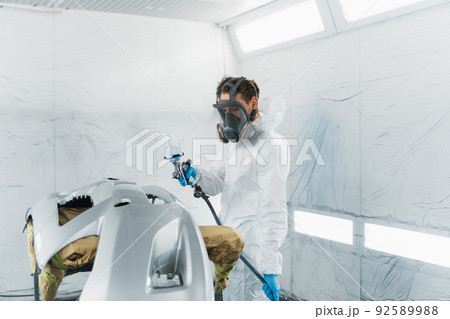Car painter in a protective suit and mask varnishes a painted bumper of a vehicle while working in a painting booth 92589988