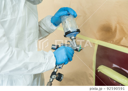 Close up view of auto painter in a protective suit and mask sprays a degreaser or other cleaner on a car door using a spray gun. Preparation of vehicle body elements for local repair Close up view of auto painter in a protective suit and mask sprays a degreaser or other cleaner on a car door using a spray gun. Preparation of vehicle body elements for local repair 92590591