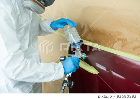 Close up view of auto painter in a protective suit and mask sprays a degreaser or other cleaner on a car door using a spray gun. Preparation of vehicle body elements for local repair Close up view of auto painter in a protective suit and mask sprays a degreaser or other cleaner on a car door using a spray gun. Preparation of vehicle body elements for local repair 92590592
