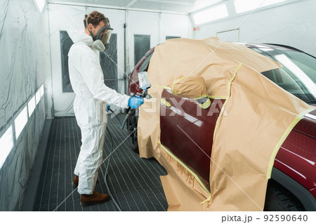 Auto painter in a protective suit and mask sprays a degreaser or other cleaner on a car door using a spray gun. Preparation of vehicle body elements for local repair Auto painter in a protective suit and mask sprays a degreaser or other cleaner on a car door using a spray gun. Preparation of vehicle body elements for local repair 92590640