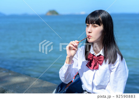 棒状のチョコレートのお菓子を綺麗な海辺の防波堤に座って食べる女子高校生　放課後　波打ち際 92590883