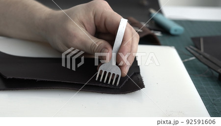 Close up men's hands make a leather product. Punching holes in the leather with a hole punch with a hammer. Tools for leather craft. 92591606