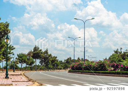 Asphalt curve empty highway, flower bushes as divider, white lines marks, lighting poles lanterns, green trees on sidewalk, blue sky cumulus clouds sun day. Beautiful summer landscape. Travel trip  92597898