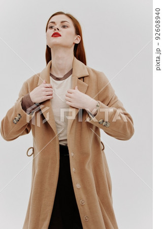 Shooting for the outerwear catalog. Red-haired beauty in a beige full-length coat in the studio on a white background 92608940
