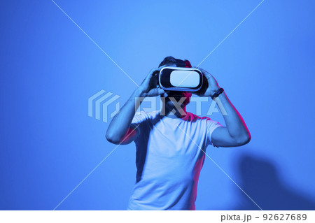 Closeup of young man in white t-shirt wearing virtual reality goggles in modern studio with a neon light. 92627689