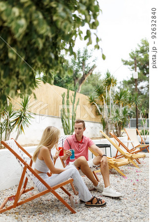 Happy young couple with glasses of lemonade sitting on deck chairs in cozy cafe place with green trees around. 92632840