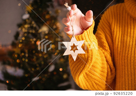 Cropped woman in orange sweater holding Xmas decoration star in front of fir tree with lights on background. Christmas holiday and New Year concept.  92635220
