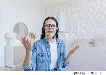 Beautiful woman looking at web camera, nervous and arguing with interlocutor, using headset for video call, working at home sitting on sofa, online conference and study 92637010