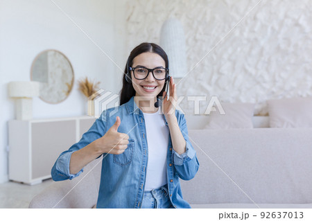 Beautiful woman working and studying at home using headset for video call, smiling and looking at camera, holding finger up, sitting on sofa in living room Beautiful woman working and studying at home using headset for video call, smiling and looking at camera, holding finger up, sitting on sofa in living room 92637013