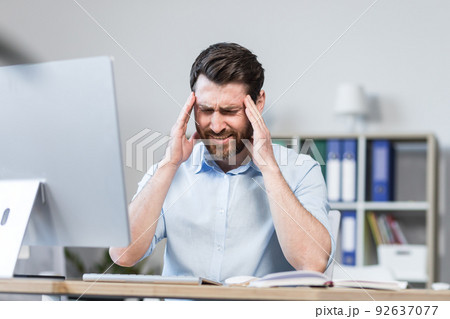 Tired and annoyed young man, office worker, manager holding his head, feeling pain, dizziness. Sitting at a computer desk in the office. 92637077