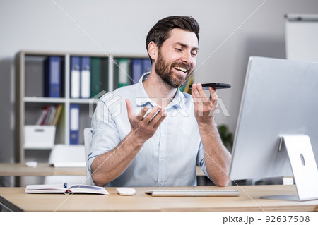 Young man with a beard, freelancer, manager, worker talking on a cell phone in the office, sitting at a computer desk, talking, waving 92637508