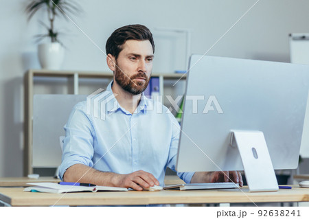 Thinking business man, working in a modern office during the day, sitting at a table, working with a computer, thinking about solving a business contract Thinking business man, working in a modern office during the day, sitting at a table, working with a computer, thinking about solving a business contract 92638241