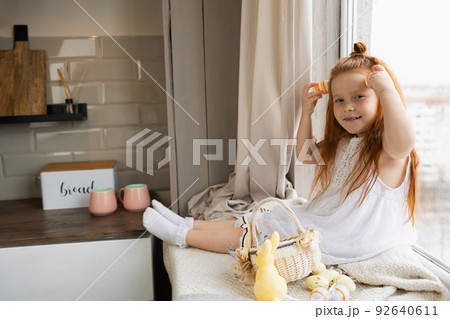 Cute girl sits on the windowsill in a cozy atmosphere and plays with Easter eggs Cute girl sits on the windowsill in a cozy atmosphere and plays with Easter eggs 92640611