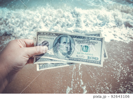 Girl holding money bill of 300 dollars on background of sea ocean waves with white foam and sand wet beach close-up. Hand wave sea ocean money dollars vacation. Concept finance money holiday traveling 92643106