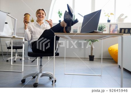 Relaxed business woman with a cup of coffee and cookies put her legs on the table Relaxed business woman with a cup of coffee and cookies put her legs on the table 92645946
