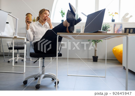 Business woman with a cup of coffee put her legs on the desk and eats cookies Business woman with a cup of coffee put her legs on the desk and eats cookies 92645947