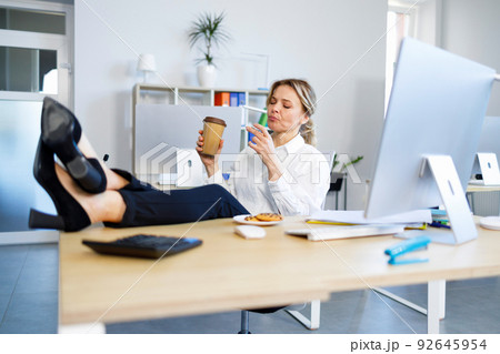 Business woman enjoying coffee and cookies while sitting in the office with her legs on the table Business woman enjoying coffee and cookies while sitting in the office with her legs on the table 92645954