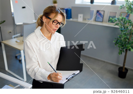 Serious business woman writing on clipboard in office Serious business woman writing on clipboard in office 92646011