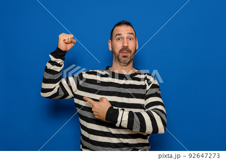 Unshaven 40 year old man in striped pullover pointing finger at biceps muscles demonstrating strength power isolated on pastel color background studio portrait. People lifestyle concept. Unshaven 40 year old man in striped pullover pointing finger at biceps muscles demonstrating strength power isolated on pastel color background studio portrait. People lifestyle concept. 92647273