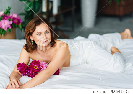 gentle brunette topless woman with a bouquet of peonies on the bed. 92649439