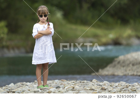 Portrait of cute pretty funny young girl with blond braids in white dress and dark sunglasses. 92650067