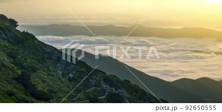 Fantastic view of mountain valley covered with low white puffy like snow clouds stretching to foggy horizon under bright morning sky with light orange glow at sunrise. Beauty of nature concept. 92650555
